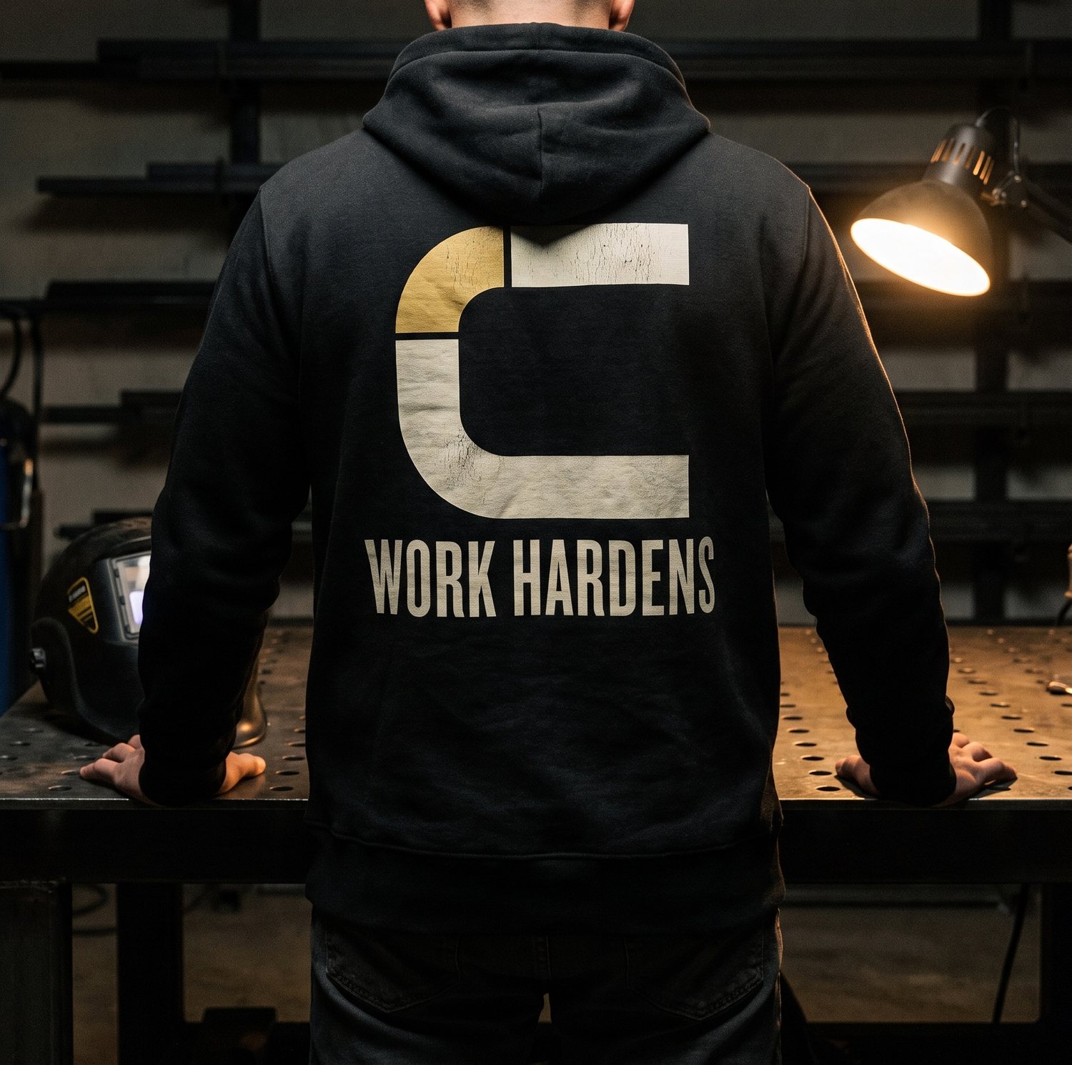 The back of a black hoodie printed with a large Coldwork mark and the words WORK HARDENS, worn at a welding table.